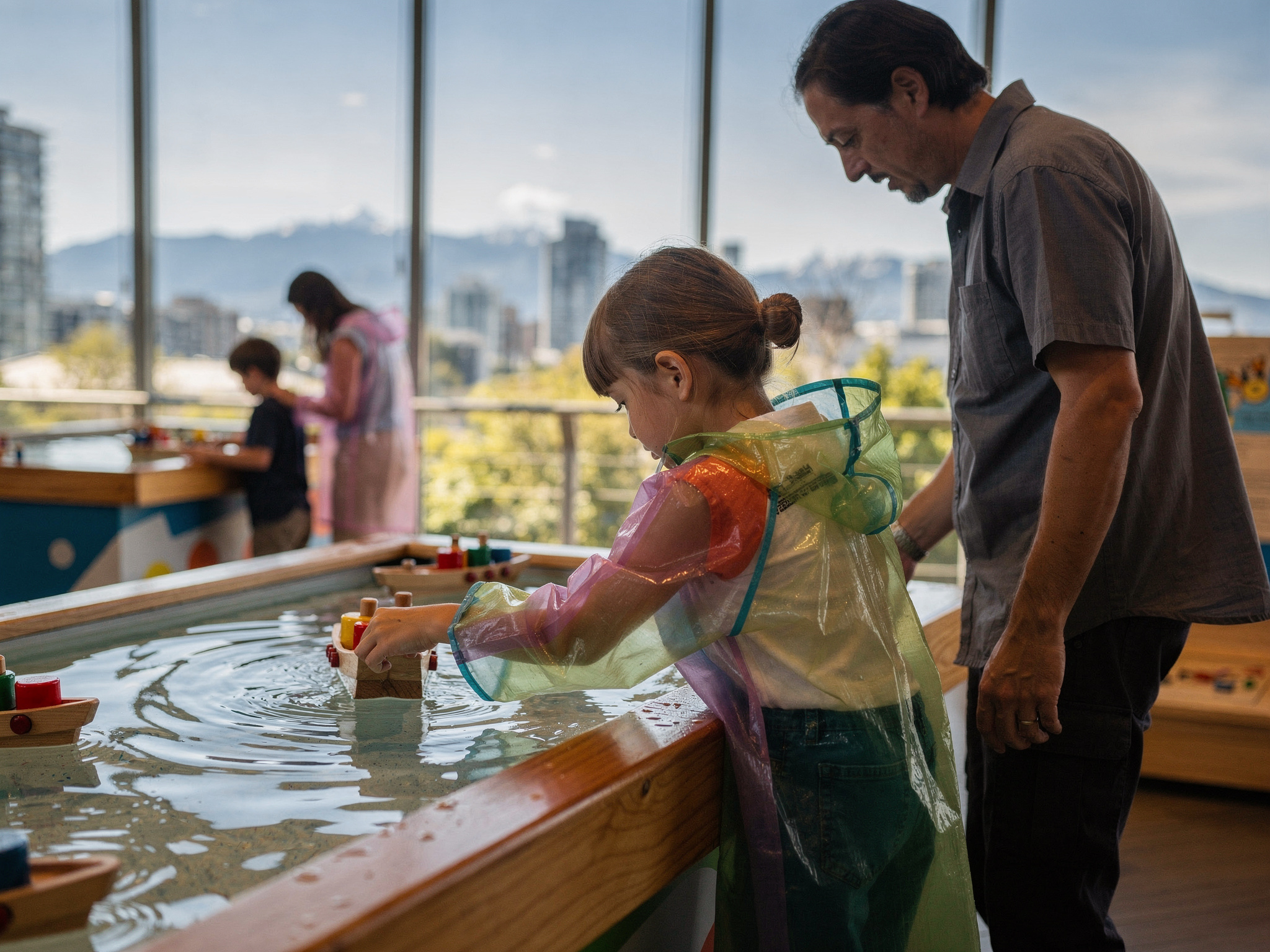 kids water play science centre v2 Educational Fun for Kids Vancouver BC A child in a rain poncho sails a toy wooden boat at a hands-on water play exhibit with a parent looking on