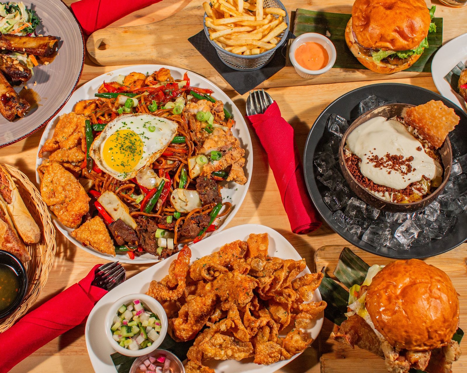 halina modern filipino food spread HALINÁ Modern Filipino Kitchen + Bar Overhead view of Halina dishes on a wooden table, including Pancit Batil Patong topped with a fried egg, crispy chicken skin, burgers with fries and a dessert set on ice